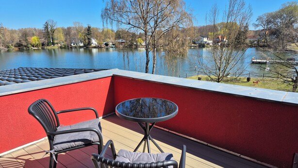 Una terraza con un muro rojo, dos sillas, una pequeña mesa redonda y vista a un lago con casas y árboles.