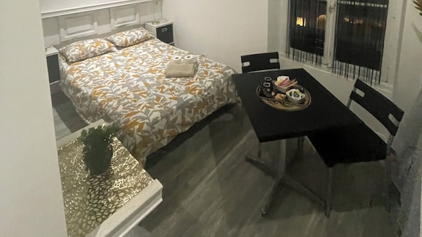 A small, well-lit bedroom with a bed, bedside tables, a window with curtains, and a dining table with chairs. - INICE  (Nice)