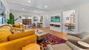 A modern living room with a sofa, coffee table, and TV. There is a dining area with chairs and a table. The room has white walls and wooden flooring. - The Wild Flower by Coastline Vacation Rentals (San Diego)