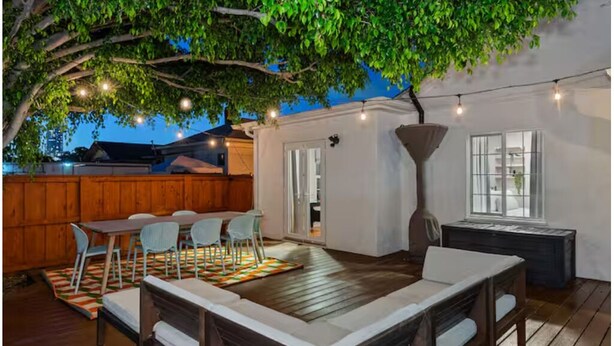 An outdoor patio with a dining table and chairs, a wooden deck, and a large tree providing shade. - The Wild Flower by Coastline Vacation Rentals (San Diego)