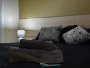 Un lit bien fait, avec des serviettes pliées et un oreiller, une lampe de chevet et un coussin à motifs sur une couvre-lit de couleur foncée.
