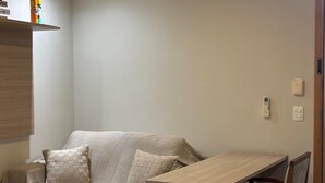 Una stanza con un divano, un tavolo in legno, una sedia, una mensola con oggetti e un telefono fissato al muro.