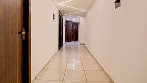 A hallway with a wooden door on the left, a wall-mounted light fixture, and a door at the end. - Deluxenana Chiloda Near Airport (Gandhinagar)
