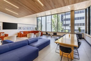 A modern interior with a large wooden ceiling, a seating area with blue and orange sofas, a long wooden table, and large windows offering a view of buildings and trees. - Boutique Studio with Pool, Gym & Walk to Midtown (Nashville)