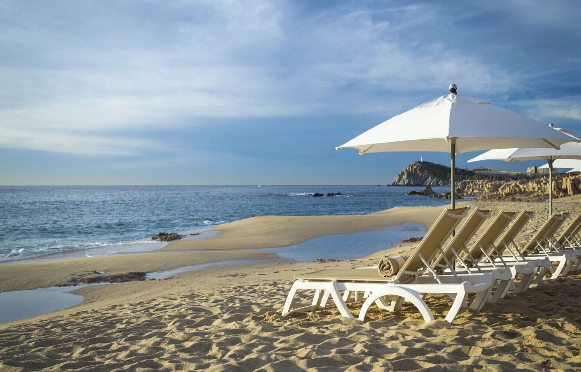 A beach with white sun loungers and umbrellas, a sandy shore, and the ocean in the background.
