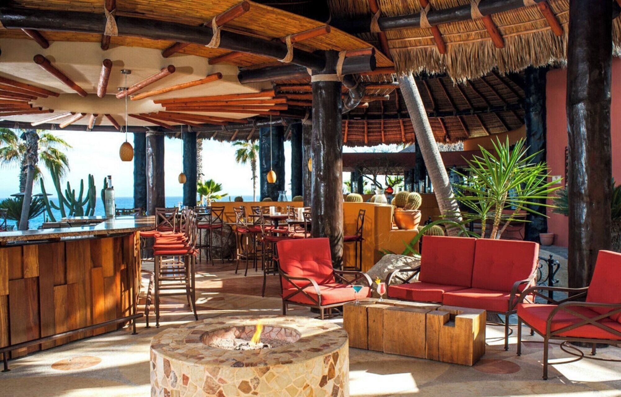 A tropical bar area with wooden furniture, a stone fire pit, and a view of the ocean.