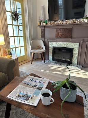 Ein Wohnzimmer mit Kamin, einer Zeitung auf dem Couchtisch, einer Zimmerpflanze und einem Fernseher.