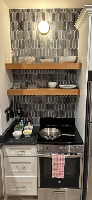Eine Küche mit einem schwarzen Herd, einer Mikrowelle und einem Edelstahlbackofen. Darüber befinden sich hölzerne Regale mit Schüsseln und Gläsern.