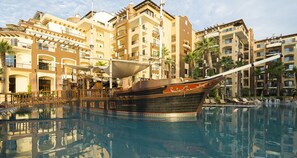 Ein großer Pool mit einer schiffsförmigen Struktur in der Mitte, umgeben von mehrstöckigen Gebäuden mit Balkonen und Palmen.