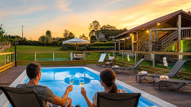A couple toasting at a poolside with a view of a stone building and a grassy area. - Casa das Xacias (A Airoá)