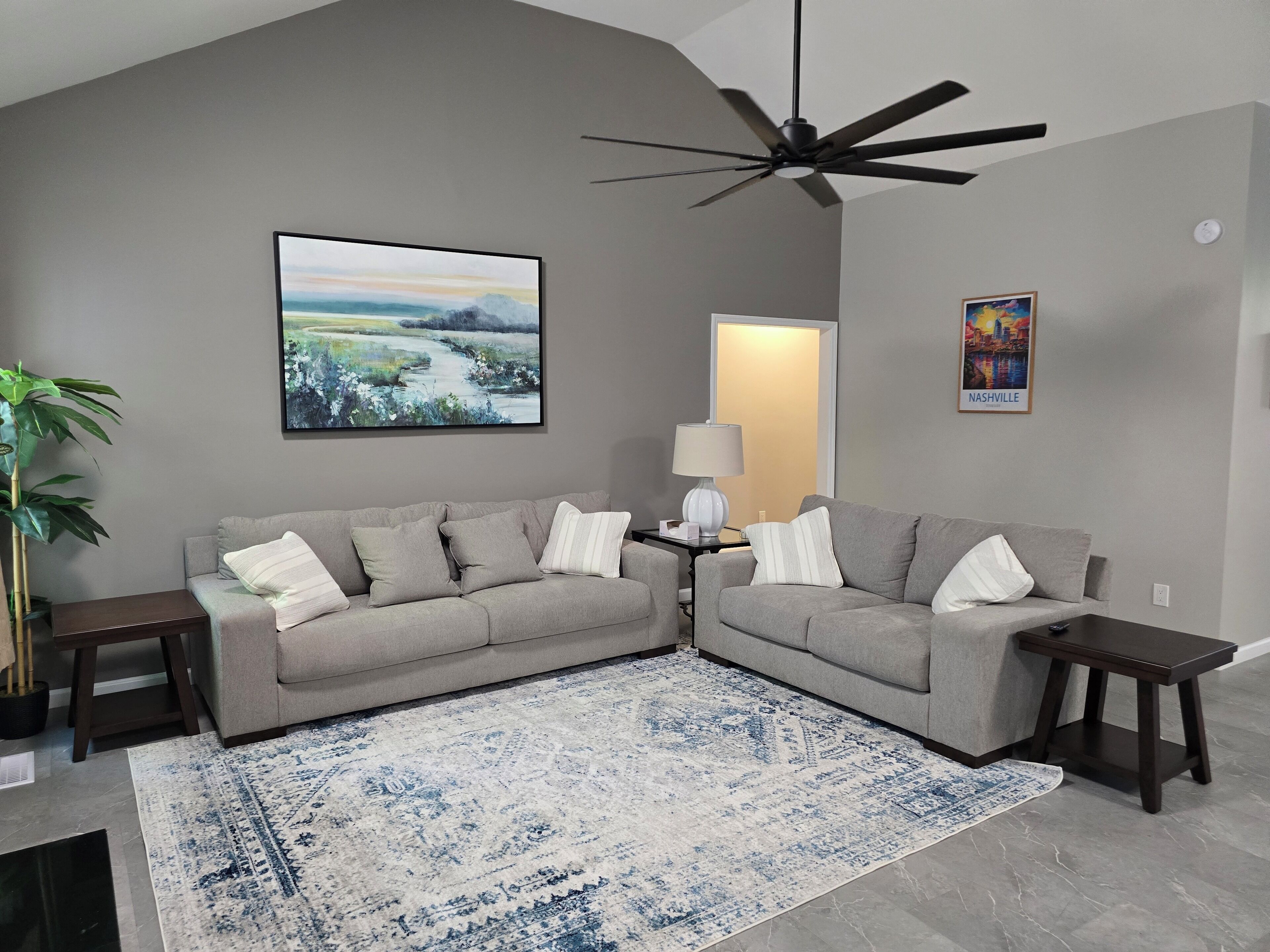Ein Wohnzimmer mit zwei grauen Sofas, einem blau-weißen Teppich, einem Deckenventilator, einem gerahmten Bild und einer Lampe.