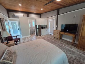 Une chambre à coucher avec un lit, un bureau, une lampe, un téléviseur posé sur un meuble en bois, et une cuisine à l’arrière-plan.