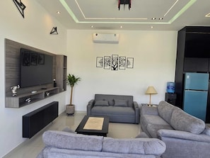 A modern living room with a flat-screen TV, grey sofas, a coffee table, and a water dispenser.