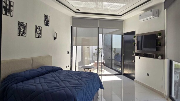 Une chambre à coucher moderne avec un grand lit, un téléviseur fixé au mur et une porte-fenêtre coulissante donnant sur un balcon.