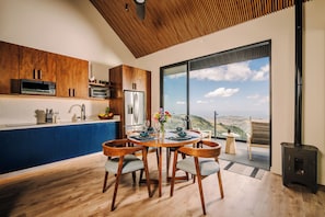 A modern kitchen with wooden cabinets, a dining table with chairs, and a large window offering a view of the hills.