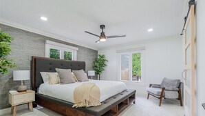Une chambre à coucher avec un grand lit, des tables de chevet, un ventilateur de plafond et une vue sur la verdure par les fenêtres.