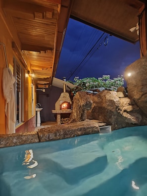 A stone fireplace with a fire, a pool, and a wooden ceiling. - Sweet Townhouse, 5 min. walk to La Fortuna centro, AC, Mini Pool w Volcano View! (La Fortuna)