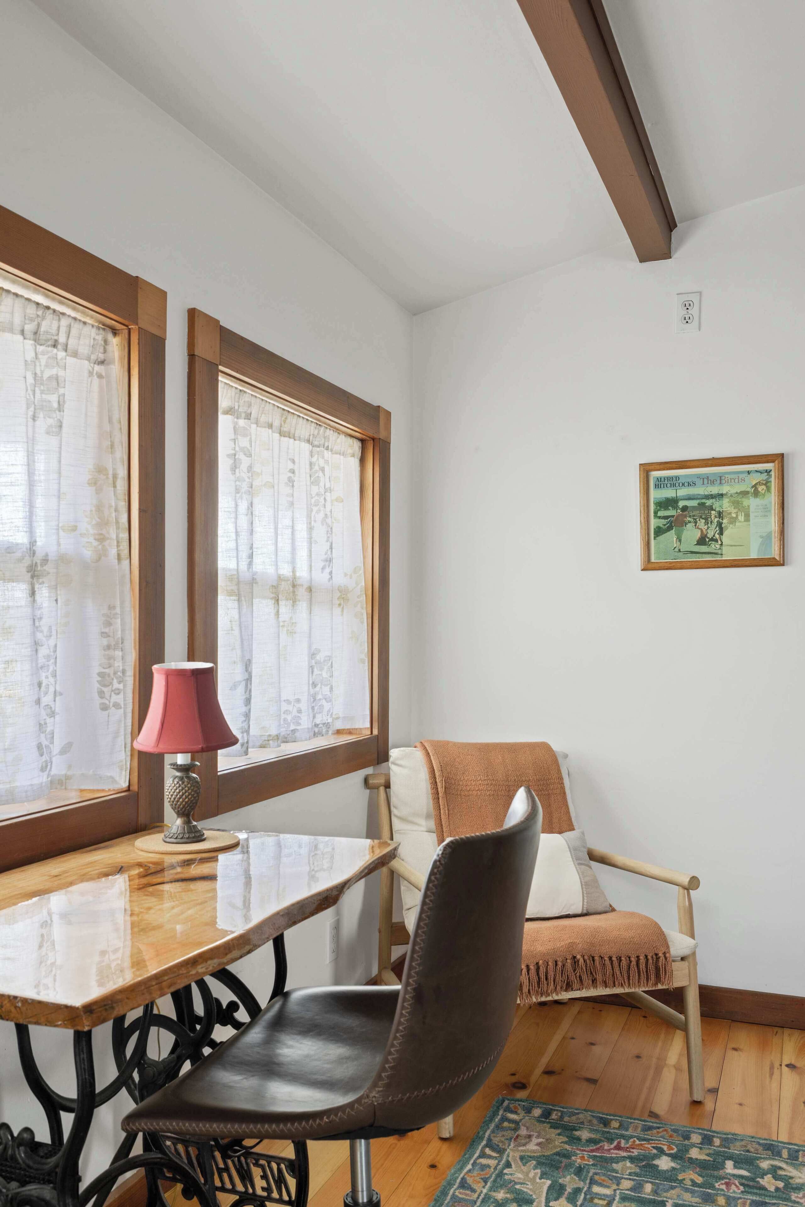 Een kamer met een houten tafel, een stoel, een lamp, een raam met gordijnen, een vloerkleed en een ingelijst beeld aan de muur.