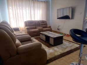 A living room with a sofa set, a coffee table, a TV mounted on the wall, and a bar stool.