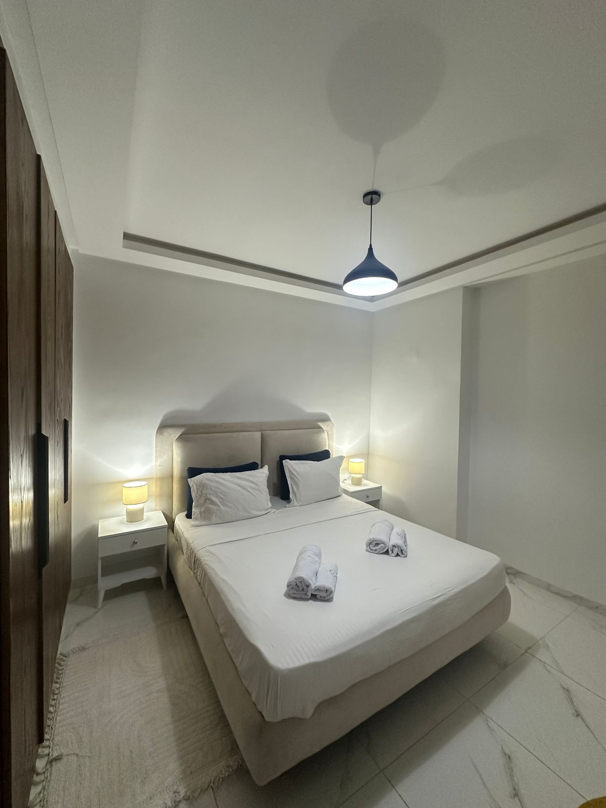 Una cama bien tendida con dos almohadas, una lámpara de noche y un aplique de techo.