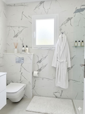 A bathroom with a toilet, a shower, a white bath mat, a towel rack with a bathrobe, and toiletries on a shelf. - Exceptional Apartment Luxury and Absolute Comfort (Tunis)