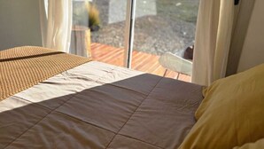 Ein Bett mit einer braunen Steude und einem gelben Kissen, eine gewebte Matte und Blick auf Berge durch ein Fenster.