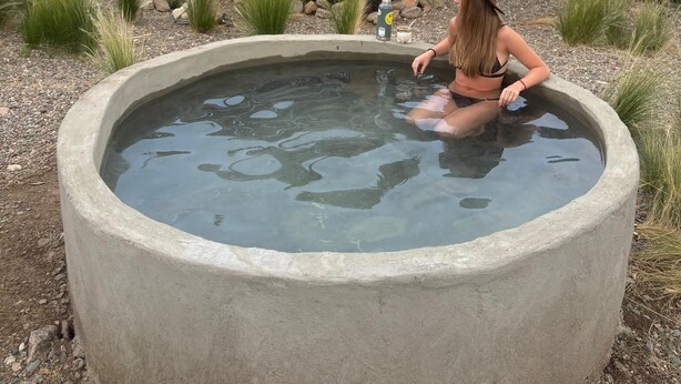 Ein Beton-Whirlpool vor einer Hütte mit einer Terrasse, Stühlen und einem Tisch.