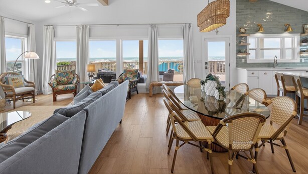 A spacious living area with a large sofa, dining table, and chairs, a balcony with a view of the ocean, and a kitchen with white cabinets. - True Beachfront New Build with Huge Oceanview Deck - Sans Souci Sleeps 12 (Port Aransas)