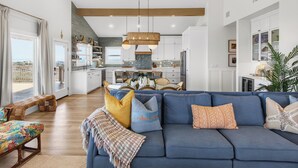 A modern living room with a blue sofa, a dining area with a table and chairs, and a kitchen with white cabinets. - True Beachfront New Build with Huge Oceanview Deck - Sans Souci Sleeps 12 (Port Aransas)