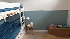 Kerrossänky, jossa on sininen patja, pieni puinen jakkara, jossa on lamppu, pyöreä puinen pöytä ja kirjahylly kirjoineen.