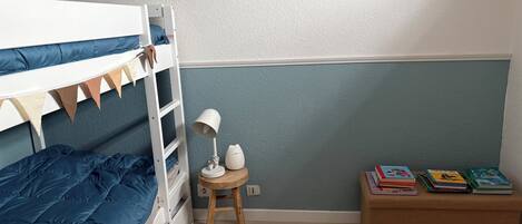 Una cama individual con colchón azul, un taburete de madera pequeño con una lámpara, una mesa de madera redonda y una estantería con libros.