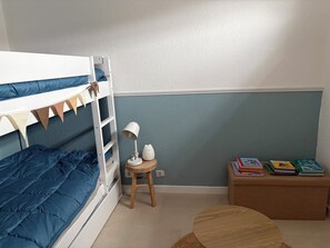 A bunk bed with a blue mattress, a small wooden stool with a lamp, a round wooden table, and a bookshelf with books. - Les Marines du Soleil in Carnon-Plage. (Mauguio)