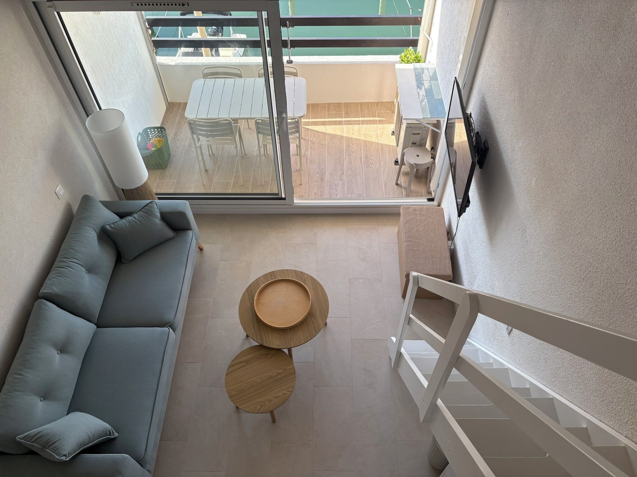 A modern interior with a grey sofa, two round wooden tables, and a view of an outdoor balcony with a table and chairs.