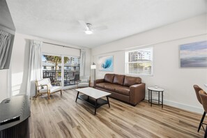 Ein modernes Wohnzimmer mit einem braunen Ledersofa, einem weißen Couchtisch, Holzboden, Deckenventilator und großen Fenstern mit Blick auf die Stadt.