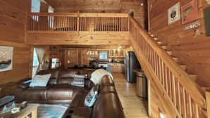 Un interior de cabaña con un sofá de cuero marrón, un comedor, una cocina y una escalera de madera.