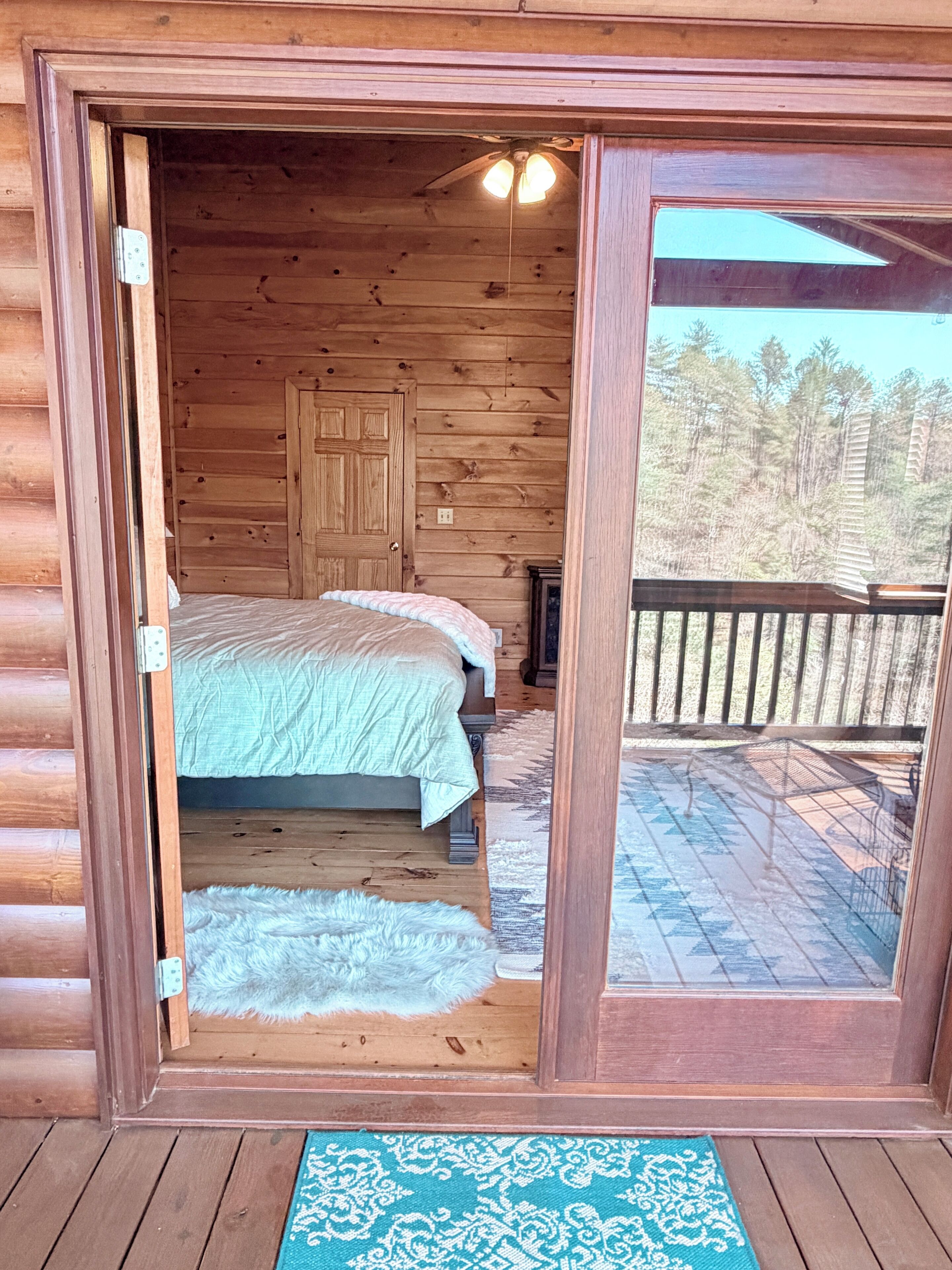 A wooden cabin bedroom with a bed, a door, and a view of a forest through a glass door.