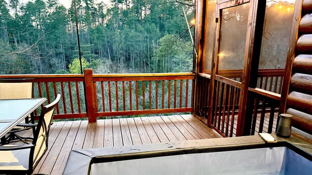 Una terraza de madera con una mesa y sillas, un jacuzzi con paredes de vidrio y vistas a un área boscosa.
