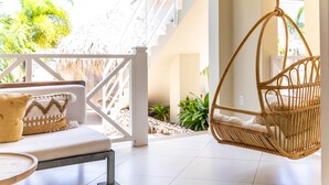 Una silla colgante de mimbre, un sofá blanco con cojines estampados y una estructura con techo de paja en un espacio amplio y luminoso con vistas a la vegetación.