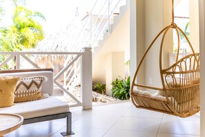 Ein Korbsessel, der an der Decke hängt, ein weißes Sofa mit gemusterten Kissen und ein strohgedecktes Gebäude in einem hellen, offenen Raum mit Blick auf Grün.
