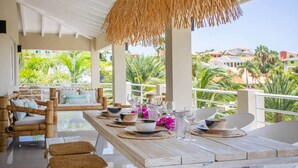 A dining area with a wooden table set for four, surrounded by wicker chairs, a thatched ceiling, and a view of palm trees and buildings. - Casa Ariba 6 person penthouse with swimming pool in JT (Jan Thiel)
