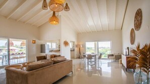 A spacious living area with a sofa, dining table, and chairs, a kitchen in the background, and large windows offering a view. - Casa Ariba 6 person penthouse with swimming pool in JT (Jan Thiel)