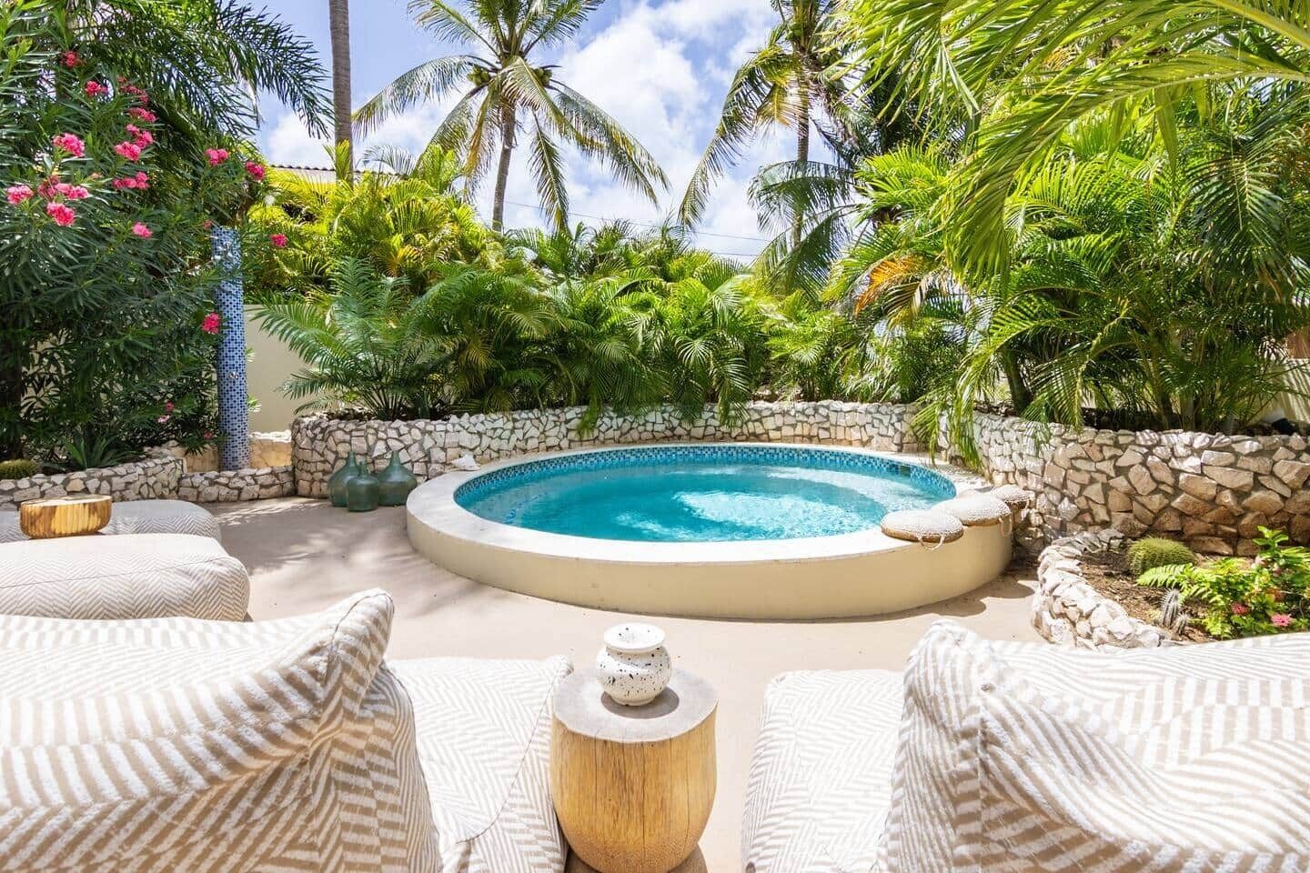 A circular hot tub surrounded by stone walls, with tropical plants and palm trees in the background.