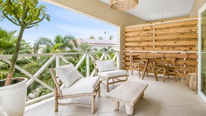 Ein Balkon mit Korbmöbeln, einer Deckenleuchte, weißen Sitzkissen, einem Holztisch und einem Blumentopf.