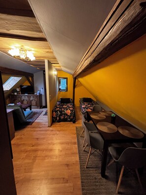 A compact room with a wooden floor, a bed with floral bedding, a small table with chairs, and a window with a view of the outside.