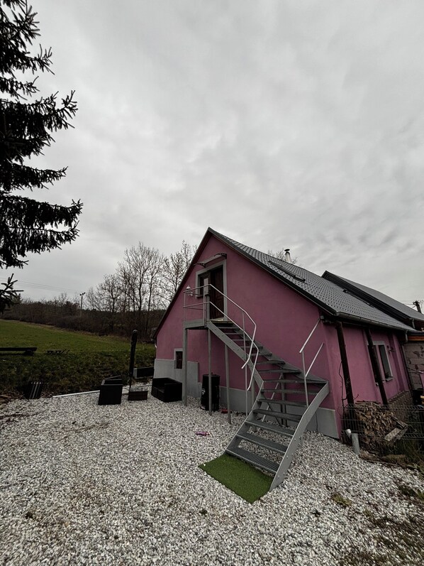 A pink building with a staircase leading to a door, gravel ground, and a tree in the background.