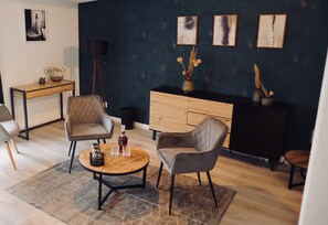 A modern living room with a dark blue accent wall, a wooden coffee table, two grey upholstered chairs, a wooden side table, and a black floor lamp.