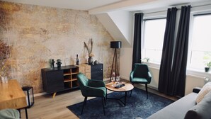 A modern living room with a sloped ceiling, a dark green sofa, two green armchairs, a small round coffee table, a dark floor lamp, and a large window with dark curtains. - Penion 44444 (Schkopau)