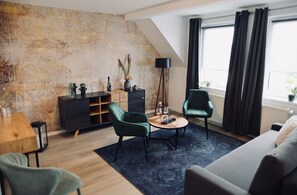 A modern living room with a sloped ceiling, a dark green sofa, two green armchairs, a small round coffee table, a dark floor lamp, and a large window with dark curtains.