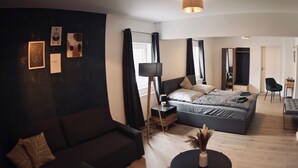 A modern bedroom with a dark-colored sofa, a round table with a vase, a large bed with multiple pillows, and a bedside table with a lamp. - Penion 44444 (Schkopau)