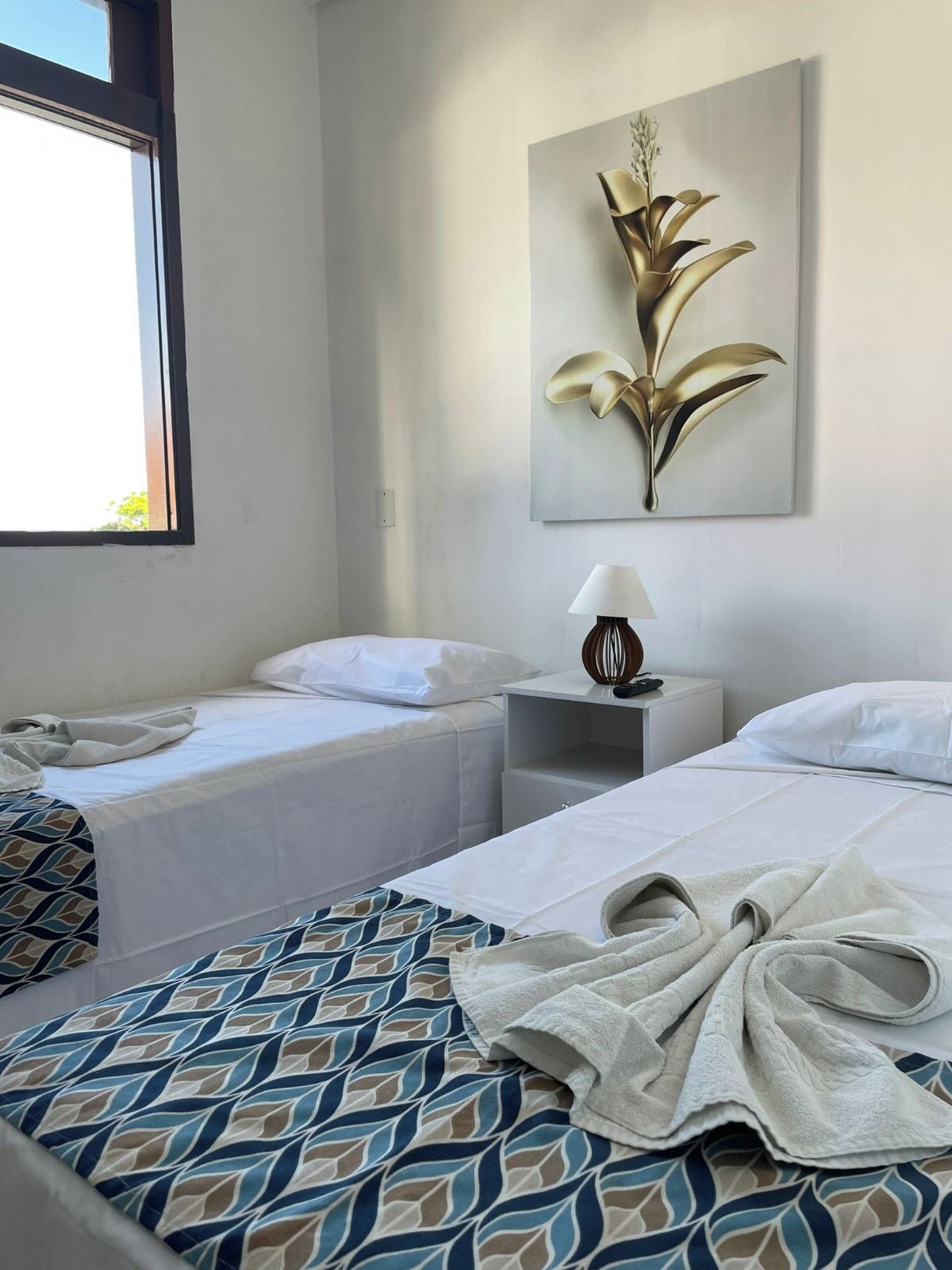 Quarto de hotel com duas camas, uma janela, mesa de cabeceira com abajur e uma obra de arte na parede.
